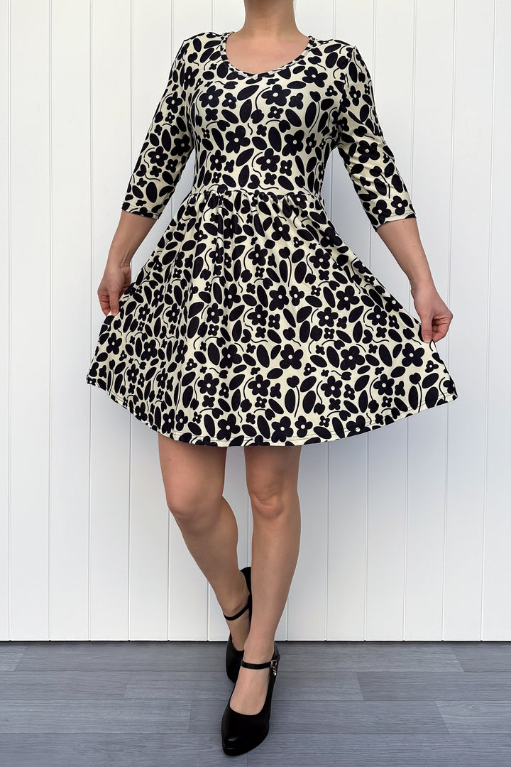 Person wearing a monochrome print dress with flowers and an ivory background, against a white wooden panel background