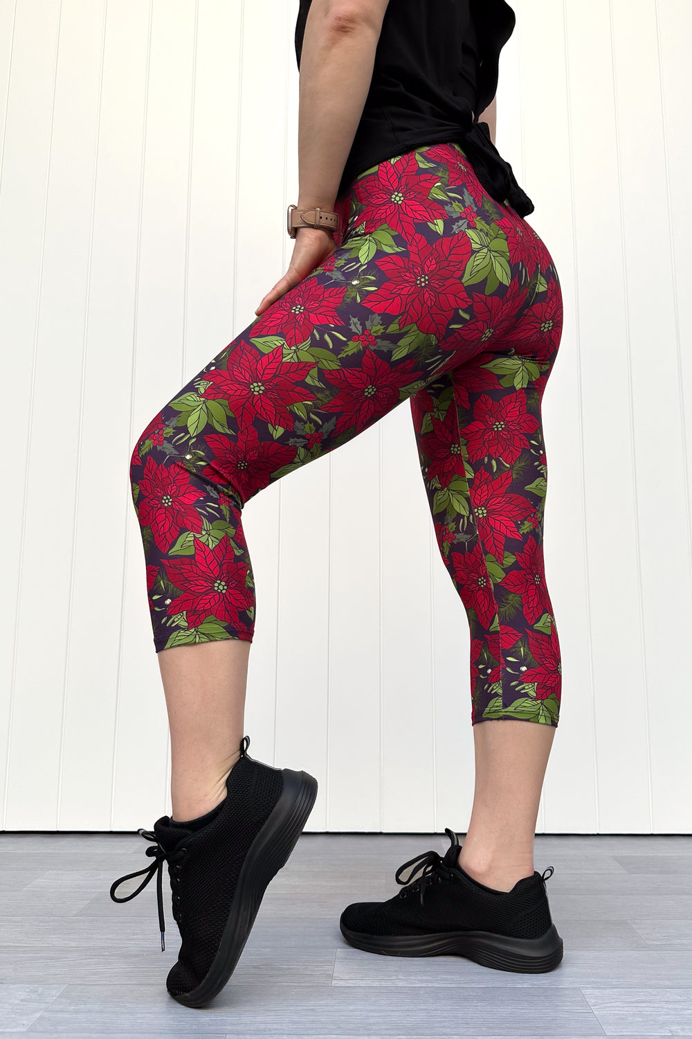 Person wearing red floral Christmas capri leggings, standing against a white wall.