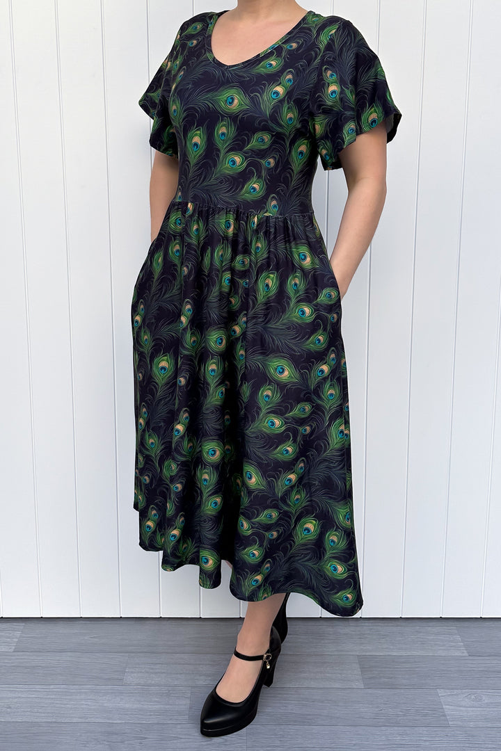 Woman wearing a black dress with peacock feather pattern against a white wooden panel background
