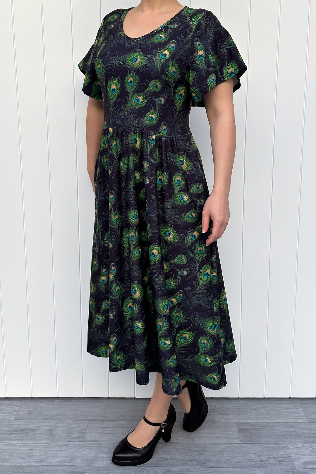 Person wearing a black dress with peacock feather pattern against a white wooden panel background