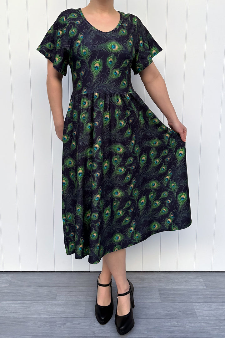 Woman wearing a black dress with peacock feather pattern against a white wooden panel background