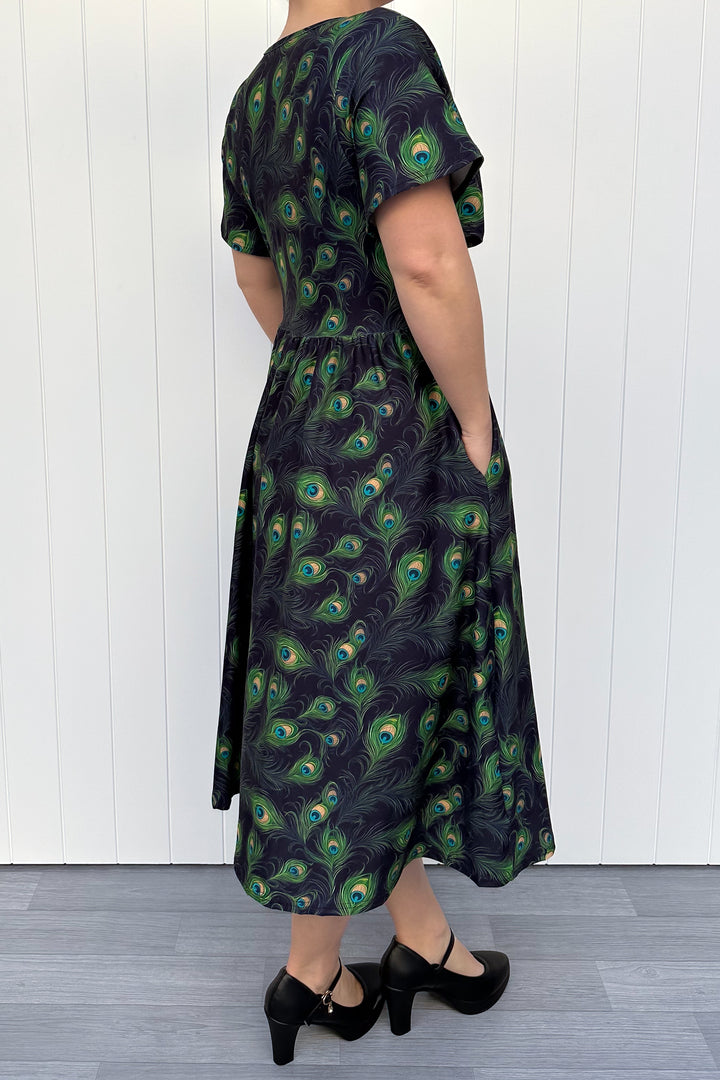 Person wearing a black dress with peacock feather pattern against a white wooden panel background