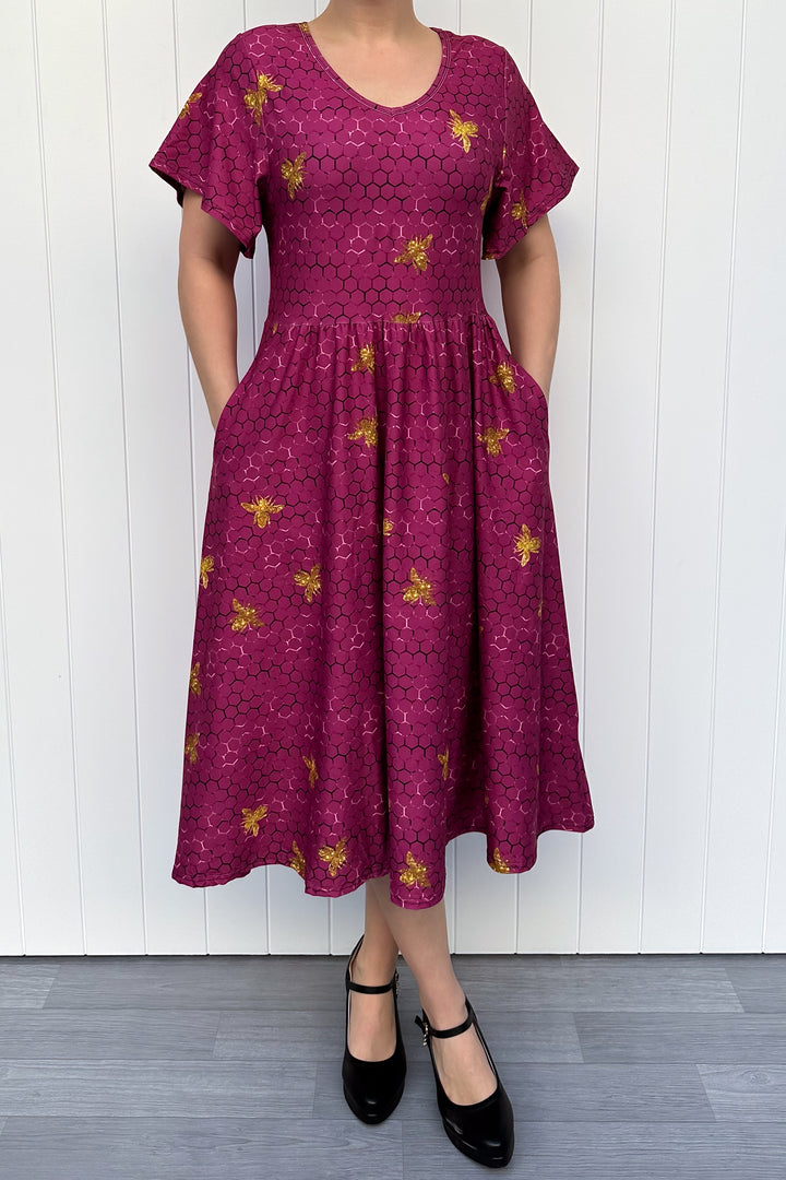 Woman wearing a magenta dress with golden bee patterns against a white wooden panel background