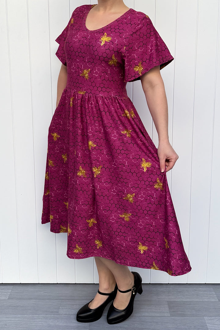 Woman wearing a magenta dress with golden bee patterns against a white wooden panel background