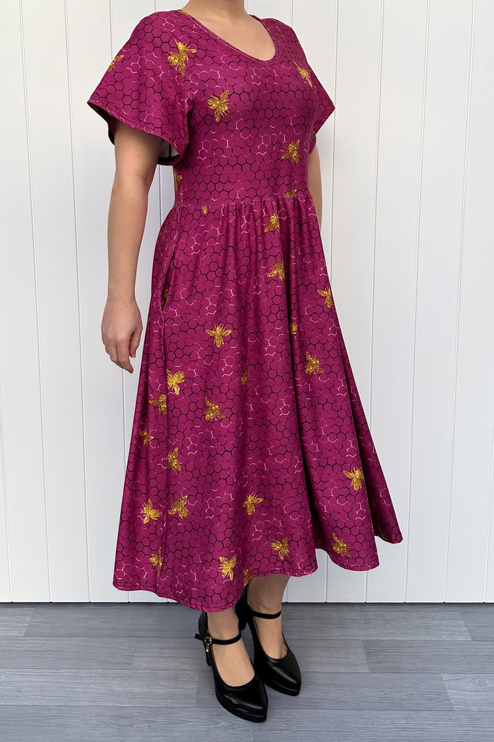 Woman wearing a magenta dress with golden bee patterns against a white wooden panel background